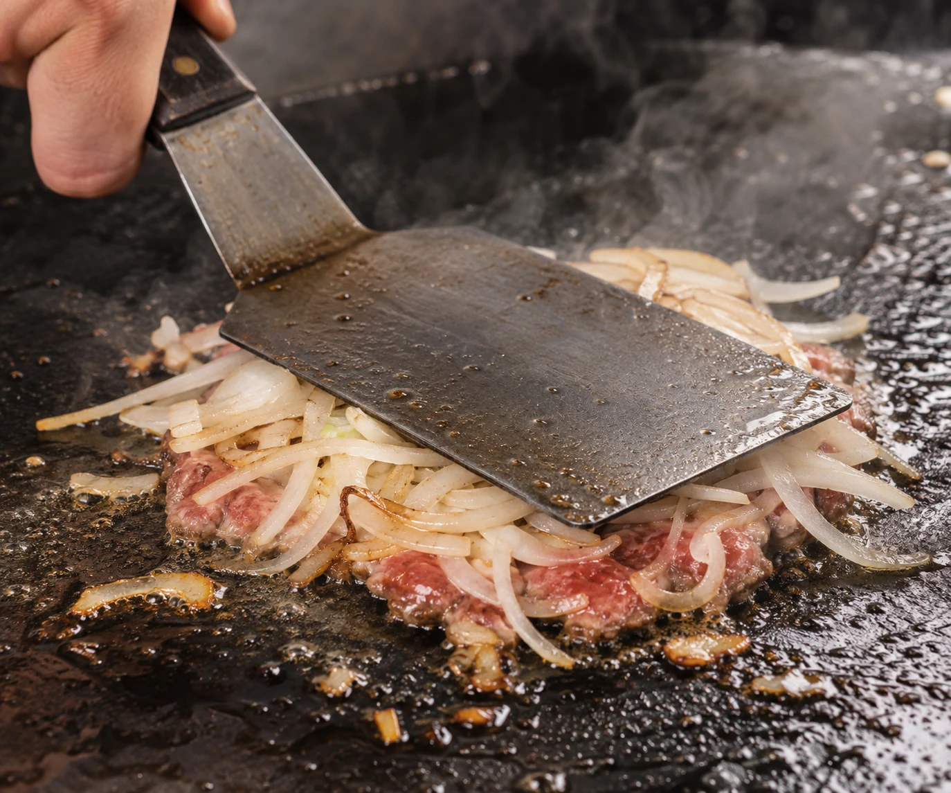 Paper-thin yellow onions are pressed into a loosely formed beef patty on cast iron.