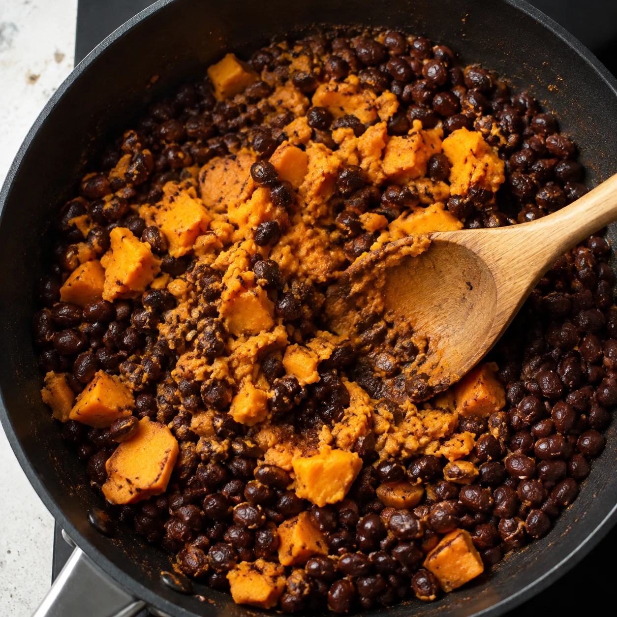 Seasoned black beans and tender sweet potato are being gently mashed together in a skillet with a wooden spoon.