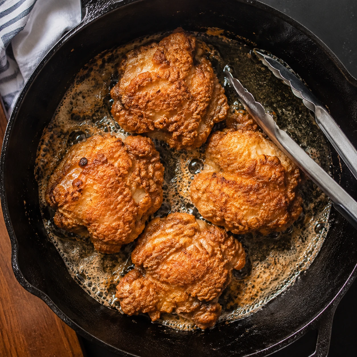 Craggy-coated chicken thighs shallow-fry in a skillet with bubbling oil and a developing golden crust.