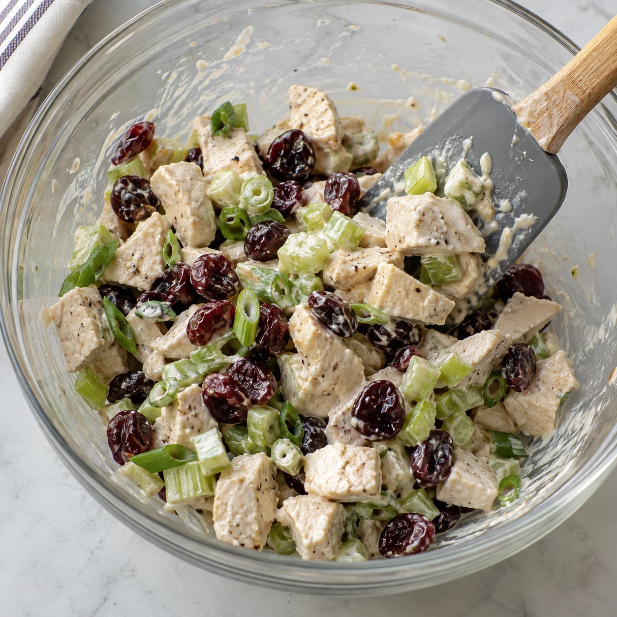 A spatula gently folds tart cherries, diced celery, scallions, and dressing through chicken pieces.