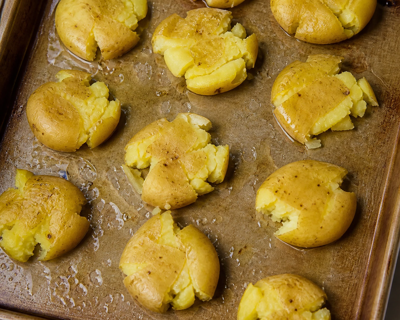 Tender potatoes are smashed on an oiled sheet pan with rough edges ready to become crisp.