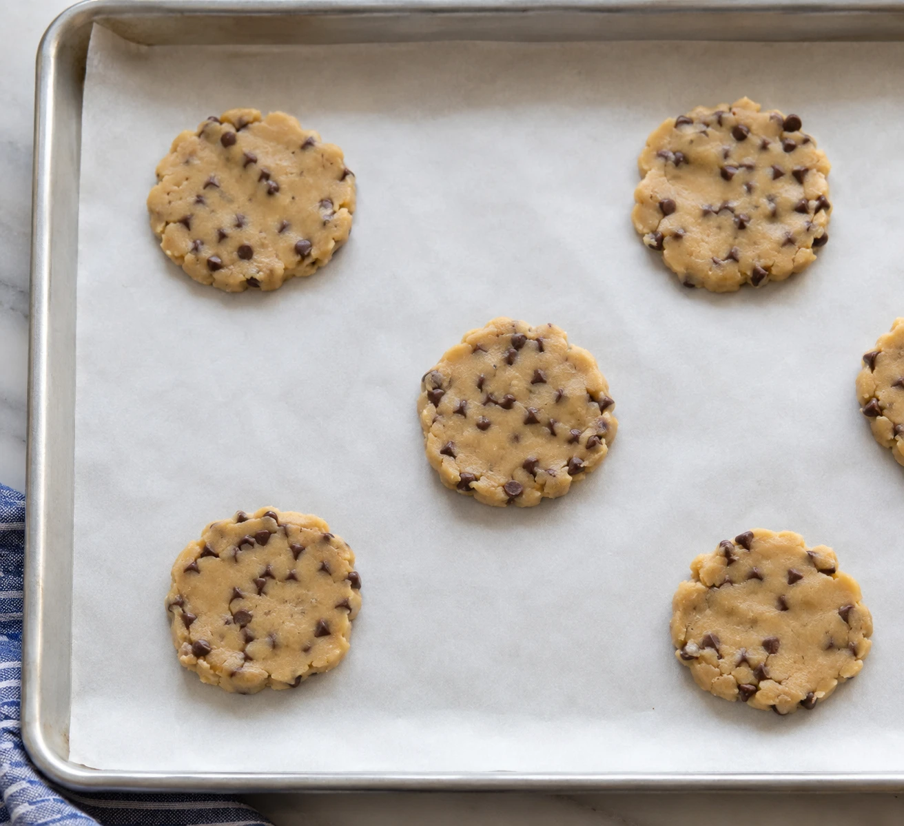 Thin disks of chocolate chip cookie dough sit widely spaced on a parchment-lined baking sheet.